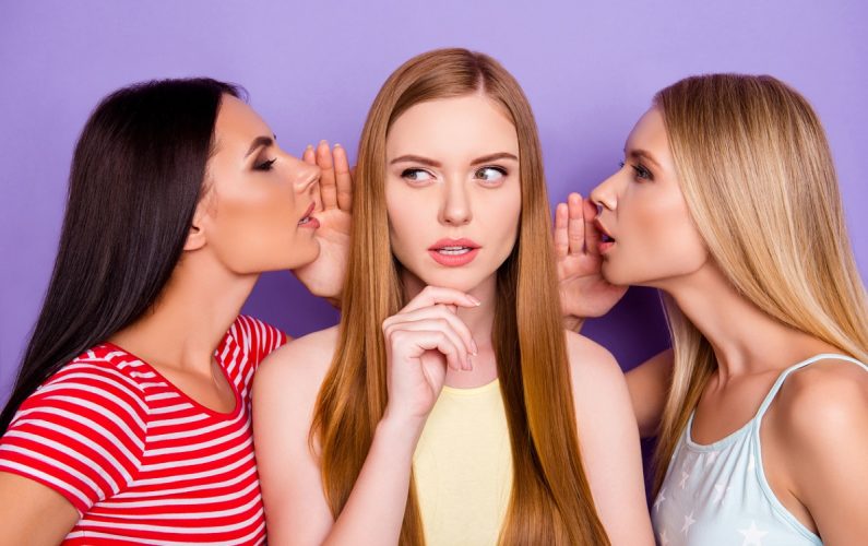 Portrait of thoughtful minded girl with long hair listening gossips from friends in two ears holding hand on chin isolated on bright violet background