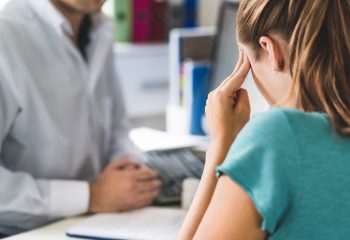 Woman suffering from bad headache or migraine. Appointment with doctor in office room. Sick and unwell lady with stress, trauma or burnout. Young patient in pain holding head with hands. Medical visit