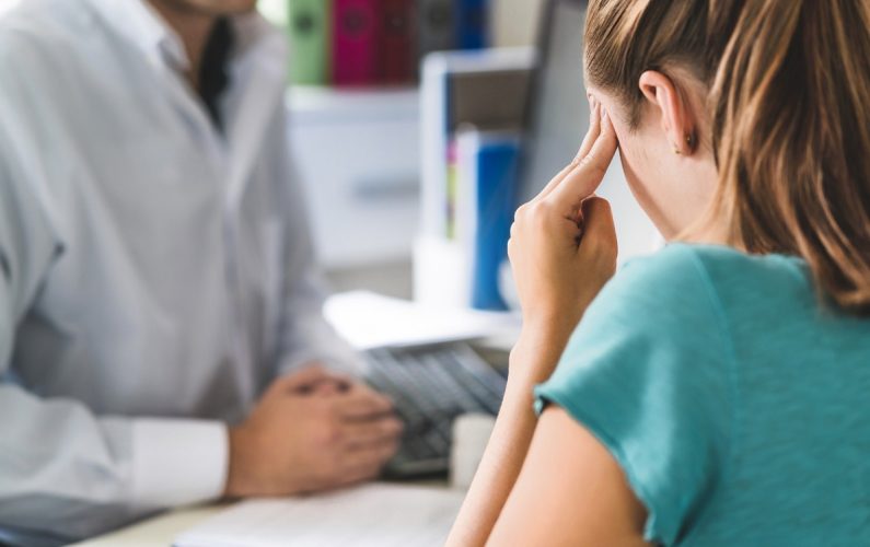 Woman suffering from bad headache or migraine. Appointment with doctor in office room. Sick and unwell lady with stress, trauma or burnout. Young patient in pain holding head with hands. Medical visit